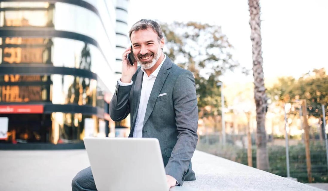 Financial services group CEO sitting on a wall with secure laptop and phone outside office buildings