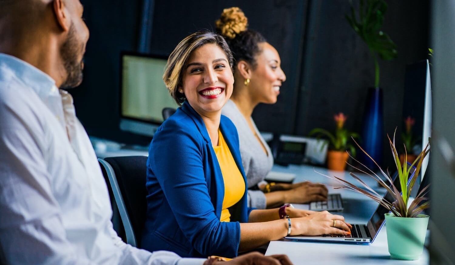 Customer service agent with colleagues at her desk smiling and sending secure email on her computer