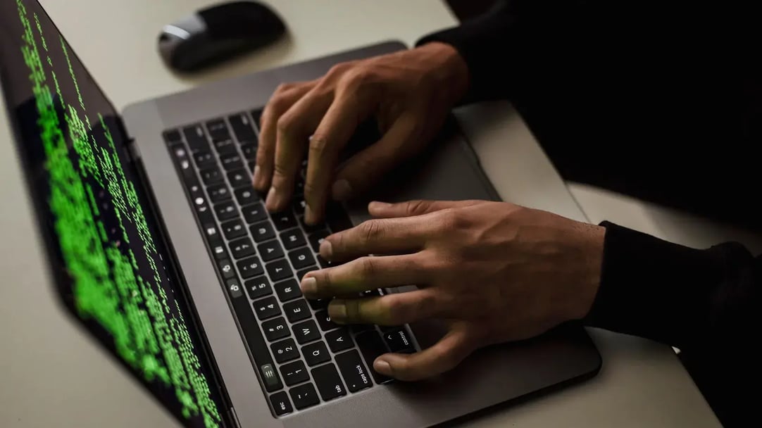 Hands typing on a laptop in a dark office, with green code cascading across the screen